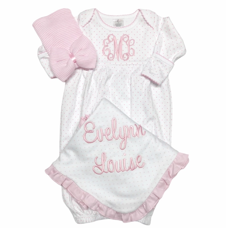 May include: A baby gift set featuring a white and pink polka dot gown, a matching hat with a bow, and a blanket. The gown has a pink embroidered monogram and trim. The blanket is embroidered with the name "Evelynn Louise".