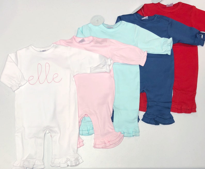 May include: Five long-sleeved baby onesies in white, pink, light blue, navy blue, and red. The white onesie has the word "elle" embroidered on the front in pink thread. All onesies have ruffled trim around the sleeves and legs.