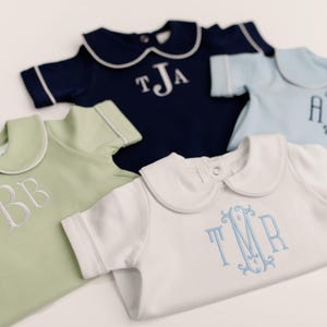 Peut inclure: Cinq barboteuses pour bébés de différentes couleurs : vert clair, bleu marine, bleu clair et blanc. Chaque barboteuse a un col Claudine et des initiales brodées. La barboteuse blanche a une broderie bleu clair.