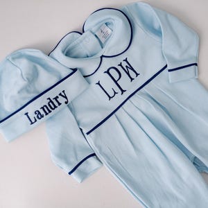 Puede incluir: Un conjunto de bebé azul claro con un gorro a juego. El conjunto tiene un cuello Peter Pan y está bordado con las iniciales "LPW". El gorro está bordado con el nombre "Landry".