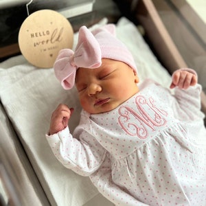May include: A newborn baby wearing a pink bow hat and a white onesie with pink polka dots and a monogram. A wooden disc with the words "Hello World" is next to the baby.