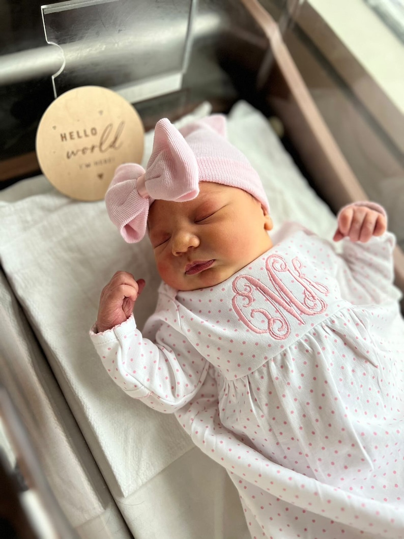 May include: A newborn baby wearing a pink bow hat and a white onesie with pink polka dots and a monogram. A wooden disc with the words "Hello World" is next to the baby.