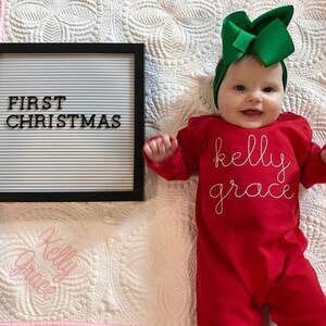 May include: A baby wearing a red long-sleeved onesie with the name "kelly grace" embroidered in white thread. The baby is wearing a green bow headband and is lying on a white quilted blanket. A black letter board with the text "FIRST CHRISTMAS" is in the background.