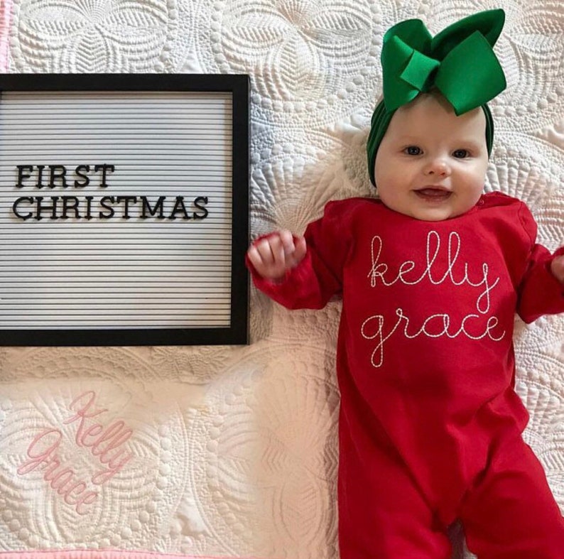 May include: A baby wearing a red long-sleeved onesie with the name "kelly grace" embroidered in white thread. The baby is wearing a green bow headband and is lying on a white quilted blanket. A black letter board with the text "FIRST CHRISTMAS" is in the background.