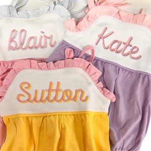 May include: Three sleeveless baby dresses with pink ruffles and embroidered names: Blair, Kate, and Sutton. The dresses are in shades of white, pink, yellow, and purple.