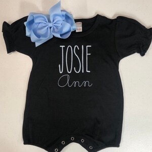 May include: Black short-sleeved baby onesie with white embroidered text that reads "JOSIE Ann". A light blue bow is on top of the onesie.
