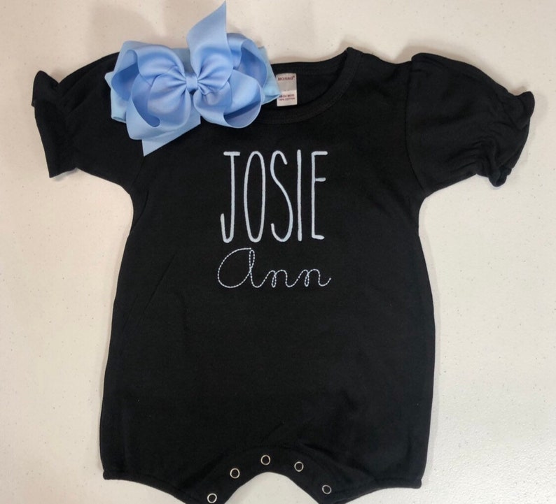 May include: Black short-sleeved baby onesie with white embroidered text that reads "JOSIE Ann". A light blue bow is on top of the onesie.