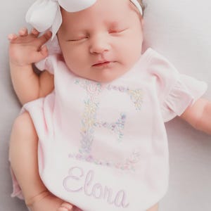 May include: A newborn baby wearing a pink onesie with the name "Elora" embroidered on it. The onesie features a floral embroidered "E" on the chest. The baby has a white bow headband.