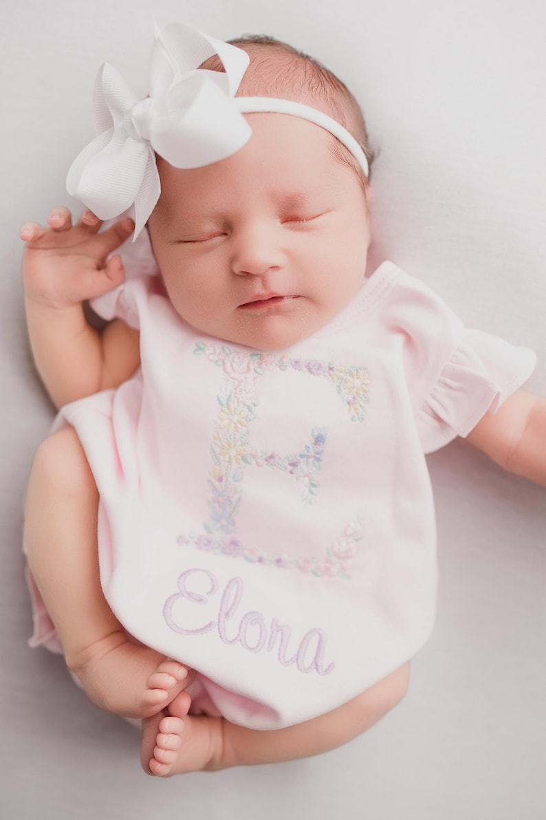 May include: A newborn baby wearing a pink onesie with the name "Elora" embroidered on it. The onesie features a floral embroidered "E" on the chest. The baby has a white bow headband.