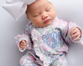 May include: A sleeping baby wearing a floral print onesie with a light blue monogram and ruffled edges. The baby is also wearing a white headband and a large white bow. The onesie has a pink, blue, and white floral pattern.