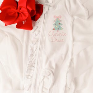 Puede incluir: Mono blanco para beb&eacute; con un dise&ntilde;o de &aacute;rbol de Navidad y el nombre "Annie Drue" bordado en rosa. Un gran lazo rojo se coloca junto al mono. El mono tiene cierre de cremallera y ribete de volantes.