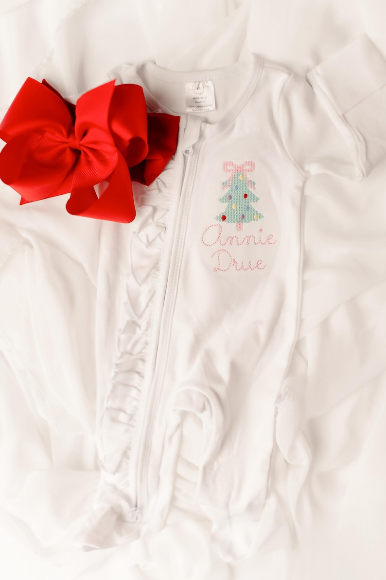 Puede incluir: Mono blanco para beb&eacute; con un dise&ntilde;o de &aacute;rbol de Navidad y el nombre "Annie Drue" bordado en rosa. Un gran lazo rojo se coloca junto al mono. El mono tiene cierre de cremallera y ribete de volantes.