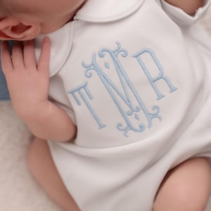 Peut inclure: Barboteuse blanche pour bébé avec col claudine et monogramme brodé bleu clair. Le monogramme présente les lettres TMR dans une police élégante et décorative. La barboteuse est douce et semble être faite d'une matière confortable.