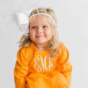 May include: A young girl wearing a bright orange dress with a white ruffle trim and a white embroidered monogram. She is wearing a white headband with a large white feather bow.
