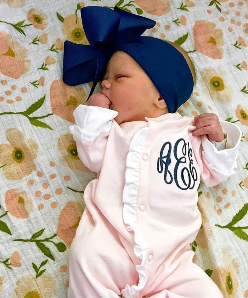 May include: A pink baby onesie with white ruffles and a navy blue monogram. The onesie is worn by a baby with a navy blue bow headband.