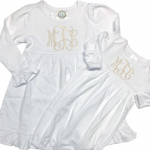 May include: Two white long-sleeved dresses with ruffled hems. The larger dress has a monogram of the letters "MJE" embroidered on the chest. The smaller dress has the same monogram embroidered on the chest.