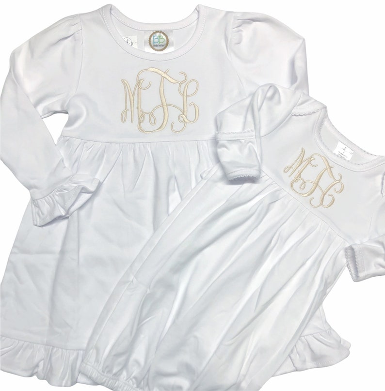 May include: Two white long-sleeved dresses with ruffled hems. The larger dress has a monogram of the letters "MJE" embroidered on the chest. The smaller dress has the same monogram embroidered on the chest.