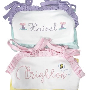 May include: Two white sleeveless dresses with embroidered names and designs. The top dress is light blue with a purple ruffle trim and a mermaid tail design. The bottom dress is yellow with a pink ruffle trim and a bee design. The names embroidered on the dresses are "Haisel" and "Brighton".
