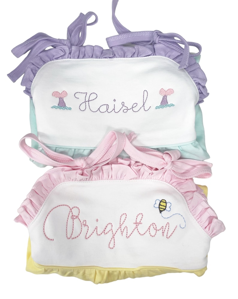 May include: Two white sleeveless dresses with embroidered names and designs. The top dress is light blue with a purple ruffle trim and a mermaid tail design. The bottom dress is yellow with a pink ruffle trim and a bee design. The names embroidered on the dresses are "Haisel" and "Brighton".