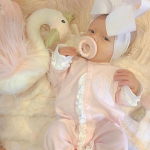 May include: A baby wearing a pink ruffled onesie with a white bow headband and a white swan plush toy. The onesie has a monogram on the front.