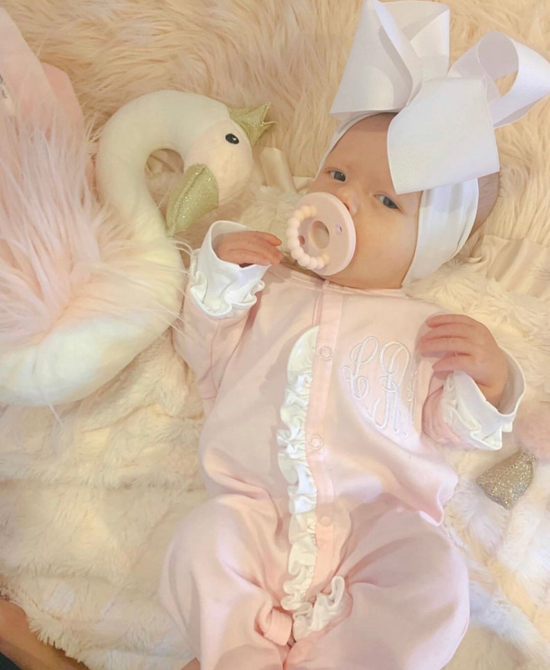 May include: A baby wearing a pink ruffled onesie with a white bow headband and a white swan plush toy. The onesie has a monogram on the front.