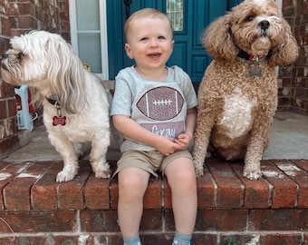 May include: A young boy wearing a grey t-shirt with a brown football graphic and the word "Gray" on it, sits on a brick stoop with two dogs. One dog is white and the other is brown. The boy is wearing white sneakers and has a big smile on his face.