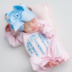 May include: A newborn baby wearing a light pink onesie with a blue floral monogram and ruffled trim. The baby is also wearing a large light blue bow and resting on a small pink pillow.