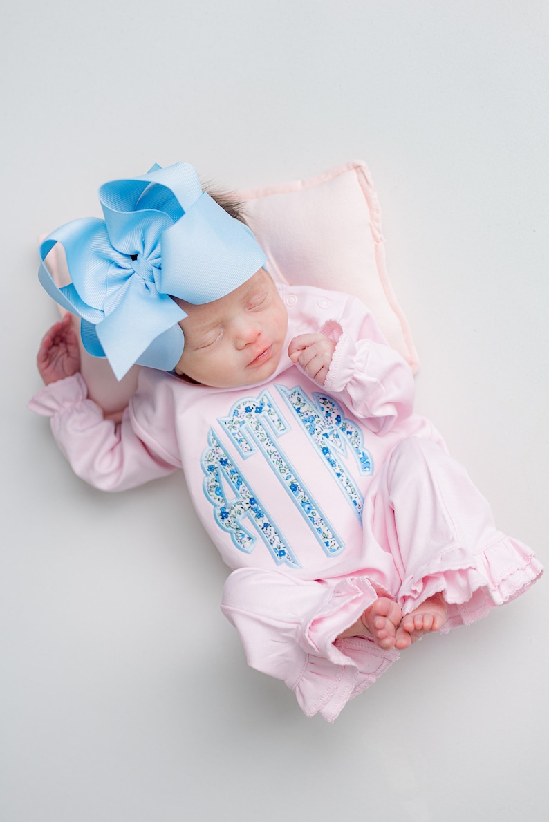 May include: A newborn baby wearing a light pink onesie with a blue floral monogram and ruffled trim. The baby is also wearing a large light blue bow and resting on a small pink pillow.