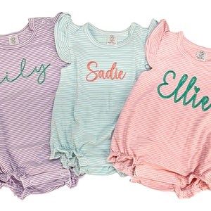 Könnte beinhalten: Drei gestreifte Baby-Bodys mit personalisierten Namen in grüner Schrift. Die Bodys sind in Lila, Hellblau und Rosa.