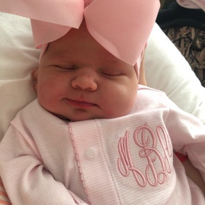 May include: A newborn baby wearing a pink and white striped onesie with a pink monogram on the front. The baby is wearing a pink bow headband.