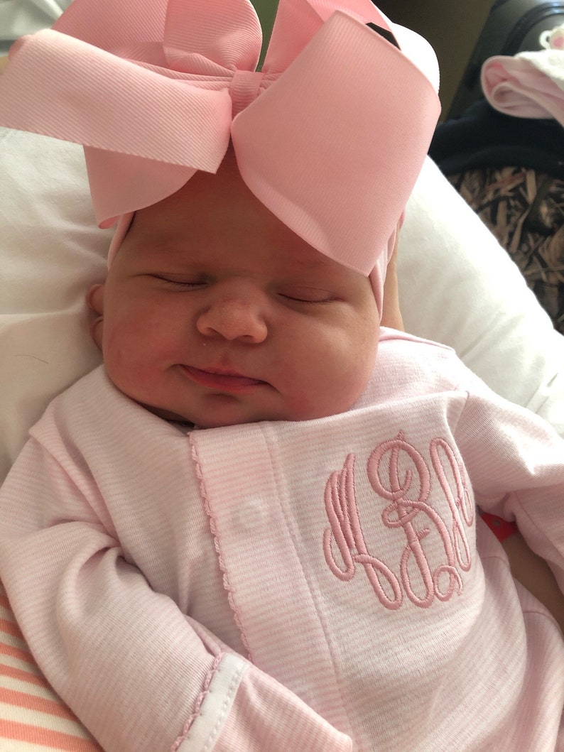 May include: A newborn baby wearing a pink and white striped onesie with a pink monogram on the front. The baby is wearing a pink bow headband.