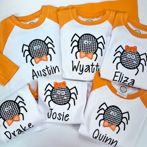 Puede incluir: Cinco camisetas de béisbol blancas y naranjas con aplicaciones de araña a cuadros negros y blancos. Cada camiseta tiene un nombre diferente bordado en la parte delantera: Austin, Wyatt, Eliza, Josie y Quinn.