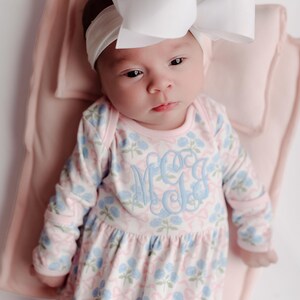 Pu&ograve; includere: Un bambino indossa una fascia bianca con un grande fiocco e un vestito a maniche lunghe con stampa floreale rosa e blu. L'abito ha un monogramma azzurro. Il bambino &egrave; sdraiato su una coperta rosa.