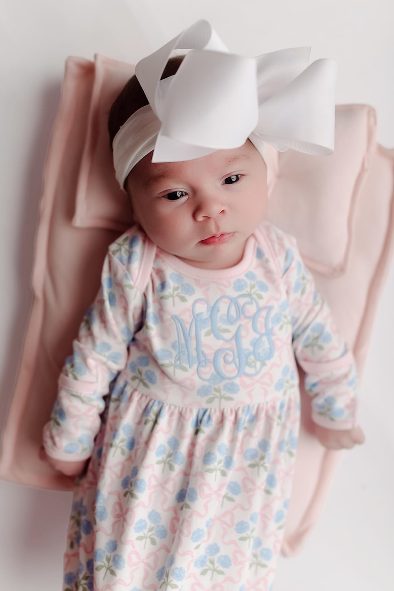 Pu&ograve; includere: Un bambino indossa una fascia bianca con un grande fiocco e un vestito a maniche lunghe con stampa floreale rosa e blu. L'abito ha un monogramma azzurro. Il bambino &egrave; sdraiato su una coperta rosa.