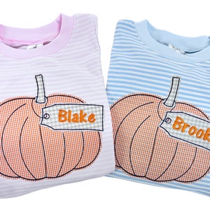 Puede incluir: Dos camisas a rayas con un dise&ntilde;o de calabaza y una etiqueta con un nombre. La camisa de la izquierda es rosa con rayas blancas y la etiqueta dice "Blake". La camisa de la derecha es azul con rayas blancas y la etiqueta dice "Brooks".