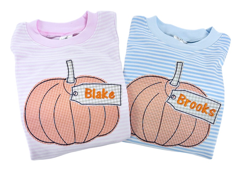 Puede incluir: Dos camisas a rayas con un dise&ntilde;o de calabaza y una etiqueta con un nombre. La camisa de la izquierda es rosa con rayas blancas y la etiqueta dice "Blake". La camisa de la derecha es azul con rayas blancas y la etiqueta dice "Brooks".