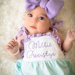 May include: A baby girl wearing a light blue and white romper with the name "Millie Annistyn" embroidered in purple. She is wearing a purple bow headband and a pearl bracelet.