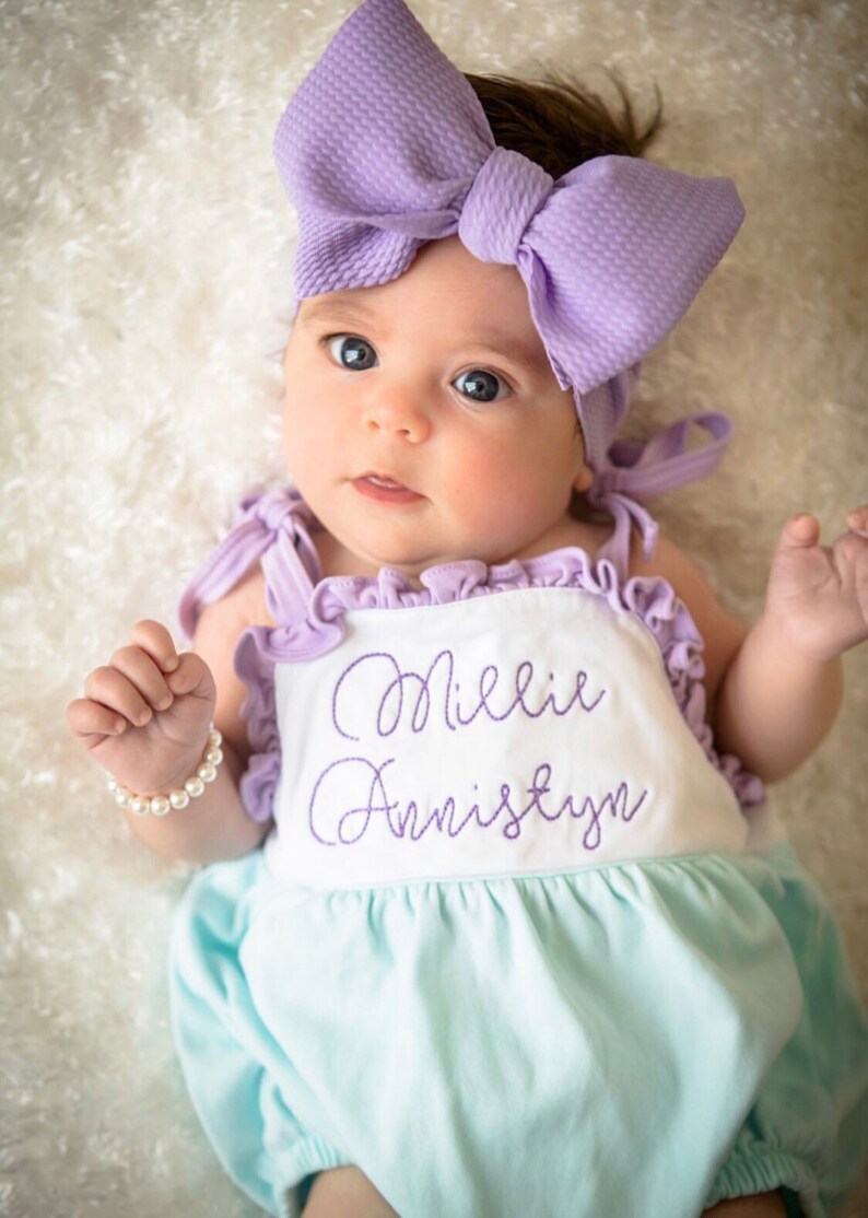 May include: A baby girl wearing a light blue and white romper with the name "Millie Annistyn" embroidered in purple. She is wearing a purple bow headband and a pearl bracelet.