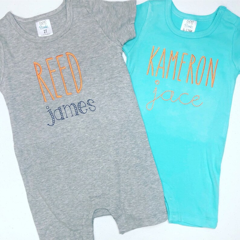 Pu&ograve; includere: Due body per neonato, uno grigio con il testo "REED james" in arancione e blu, e uno turchese con il testo "KAMERON jace" in arancione.