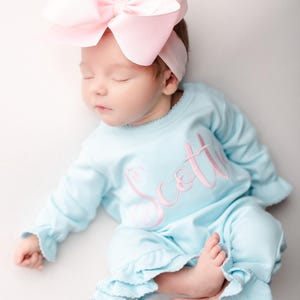 May include: A sleeping infant wearing a light blue outfit with the name "Scott" embroidered in pink. The outfit has ruffled edges. The baby is also wearing a large pink bow headband.