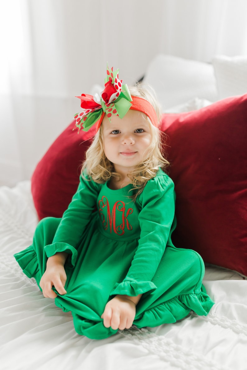 May include: A young girl wearing a green dress with a red embroidered monogram and a red headband with a red and green polka dot bow. She is sitting on a white bed with a red pillow behind her.