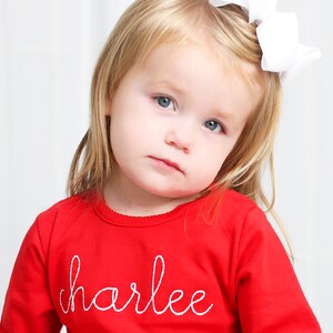May include: A young girl wearing a red long-sleeved shirt with the name "charlee" embroidered in white on the front. She has blonde hair and is wearing a white bow.