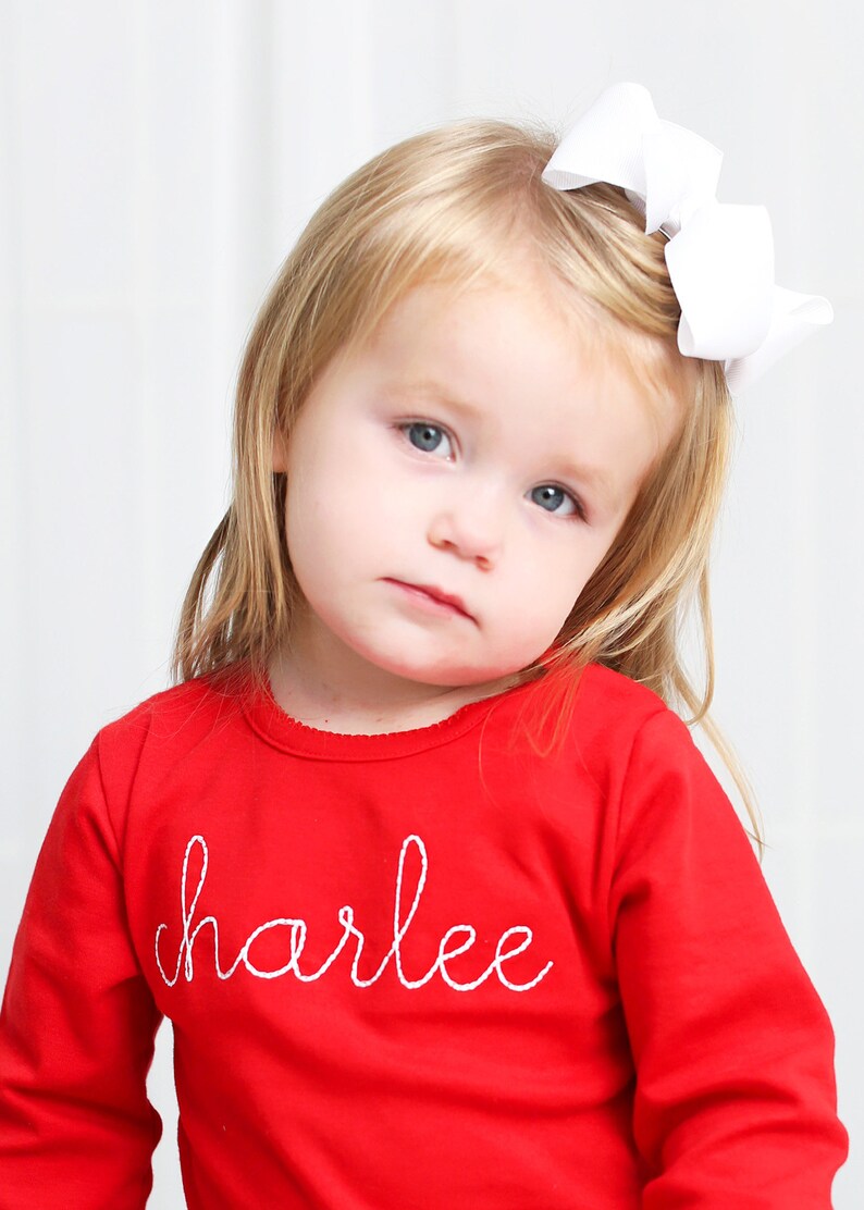 May include: A young girl wearing a red long-sleeved shirt with the name "charlee" embroidered in white on the front. She has blonde hair and is wearing a white bow.