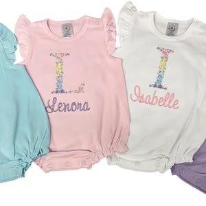 May include: Four baby rompers in pastel colors: light blue, pink, white, and lavender. Each romper features a floral initial and name embroidery. The rompers have ruffled sleeves and a bubble bottom.