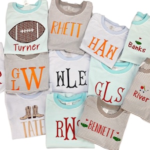 Puede incluir: Una colección de camisas a rayas con varios diseños bordados, incluyendo un balón de fútbol, un palo de golf, una bota de vaquero y iniciales. Las camisas son en tonos de azul, verde y gris.
