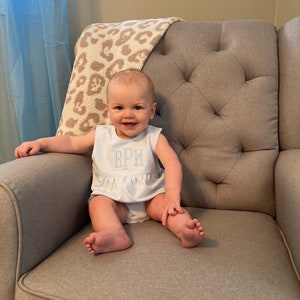 May include: A baby wearing a light blue and white striped romper with the initials "RPW" embroidered on the chest. The baby is sitting on a gray upholstered armchair with a leopard print blanket draped over the back.