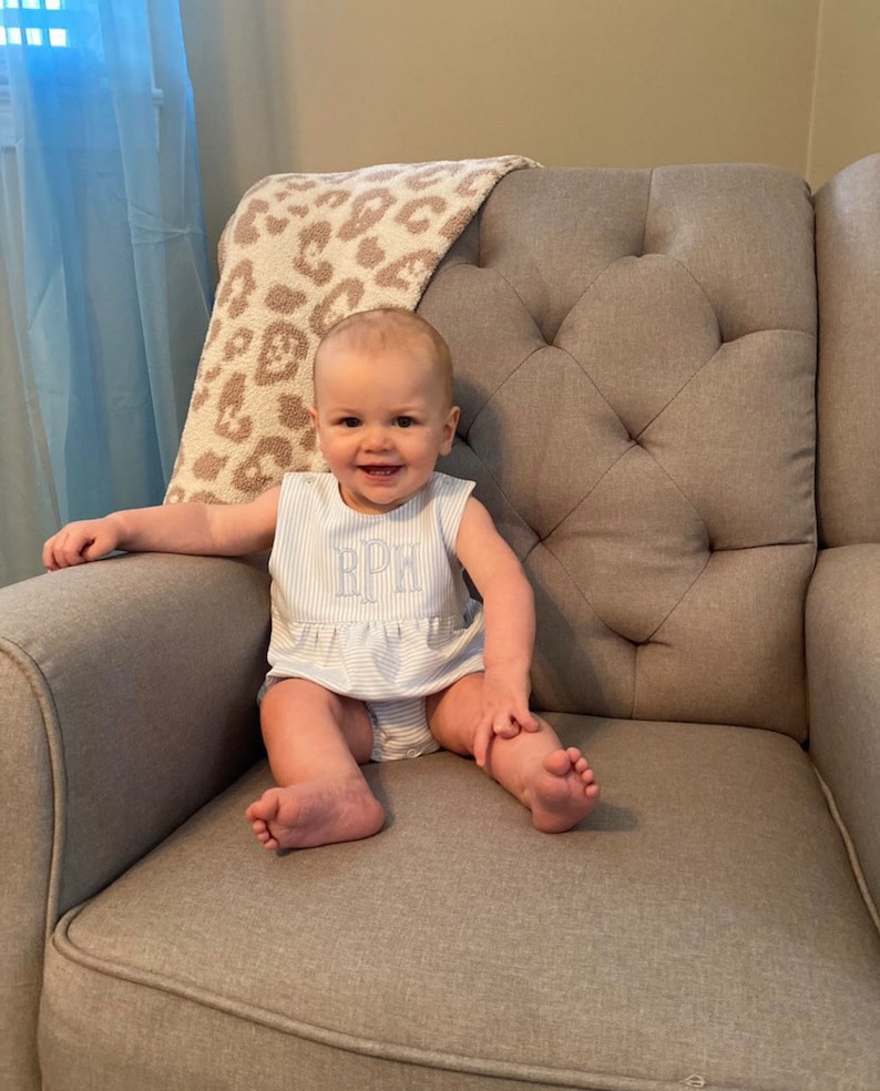May include: A baby wearing a light blue and white striped romper with the initials "RPW" embroidered on the chest. The baby is sitting on a gray upholstered armchair with a leopard print blanket draped over the back.