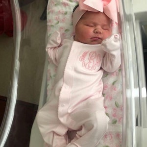 May include: A newborn baby girl wearing a pink onesie with a monogram on the front. The baby is sleeping in a clear plastic bassinet with a floral patterned blanket.