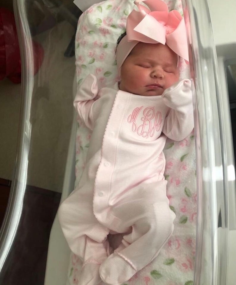 May include: A newborn baby girl wearing a pink onesie with a monogram on the front. The baby is sleeping in a clear plastic bassinet with a floral patterned blanket.