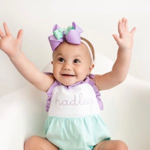 May include: A baby girl wearing a light green and white romper with a purple ruffle trim and the name "hadley" embroidered on the front. She is wearing a purple and green bow headband and has her arms raised in the air.